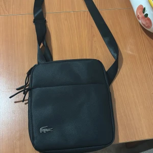 Svart axelväska från Lacoste - Snygg svart axelväska från Lacoste med klassisk krokodillogga i metall framtill. Väskan har justerbar rem, dragkedja upptill och flera fack, både invändigt och utvändigt. Tillverkad i slitstark syntet med stilren, fyrkantig form. Perfekt för att bära dina allt du behöver. Perfekt för att ge outfiten en lyxig och elegant touch🔥🔥. Tveka inte på att ställa frågor! Pris är ej hugget i sten