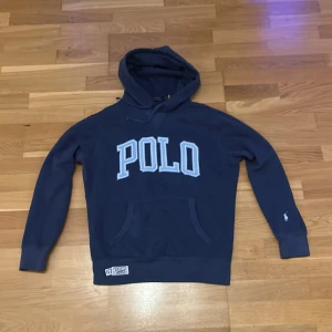 Marinblå hoodie från Polo Ralph Lauren - Snygg marinblå hoodie från Polo Ralph Lauren med stor POLO-text i ljusblått framtill och klassisk broderad logga på ärmen. Tröjan har huva med snörning och känguruficka. Tillverkad i mjuk bomull, perfekt för chill dagar.