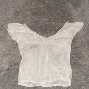 Vit blus med spetsdetalj - Supersöt vit blus med v-ringning och spetskant runt halsen. Lätt och luftig känsla, korta ärmar med lite puff och resår. Perfekt att styla med jeans eller kjol för en fräsch look.