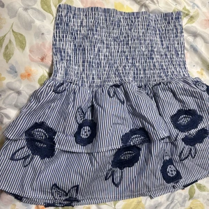 Blåvit randig kjol med broderi - Supersöt kortkjol med blå och vita ränder, volanger och mörkblå broderade blommor. Kjolen har smock i midjan för en bekväm passform och kan användas som en bandeau topp!!!