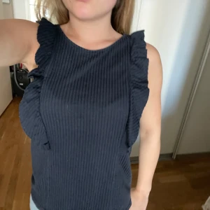 Mörkblå ribbad linnetopp med volang - Säljer en mörblå ärmlös linnetopp med ribbad struktur och volangdetaljer längs axlarna. Toppen har rund halsringning och sitter snyggt mot kroppen.🥰