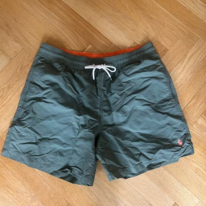 Gröna badshorts Polo Ralph Lauren M - Snygga gröna badshorts från Polo Ralph Lauren med orange midjeresår och vit snörning. Klassisk passform, meshfoder och bakficka med knapp. Liten broderad logga i rött framtill. Perfekta för stranden eller poolen! (Lite slitna vid fickan se bild 4) 