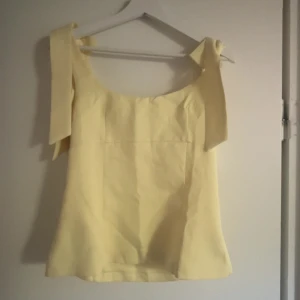 Gul linnetopp med knytband - Söt ljusgul linnetopp med breda axelband som knyts till rosetter på axlarna. Toppen har en rak passform och ärmlös. Enkel och stilren look som passar till jeans eller kjol. Säljer då den var förstor den är helt ny och har bara testat den. Kan tänka mig att sänka lite i pris