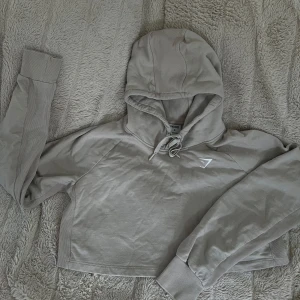 Beige croppad hoodie från Gymshark - Snygg beige croppad hoodie från Gymshark med huva och snörning. Tröjan har långa ärmar med breda muddar. supersnygg men tar plats i garderoben. 