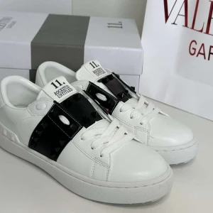 Valentino Garavani Skor - Snygga vita Valentino Rockstud Untitled sneakers med svart band och ikoniska nitar över vristen. Skorna är i skinn med rund tå, platt sula och vita snören. Perfekt för dig som vill sticka ut med en lyxig och edgy detalj i din outfit.