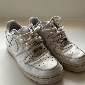 Nike Air Force 1 vita sneakers - Klassiska Nike Air Force 1 sneakers i helvitt skinn med perforerad tå, snörning och ikonisk swoosh på sidorna. Låg modell med platt sula och broderad Nike Air-logga på hälen. Perfekta för dig som gillar streetstyle och tidlös design.