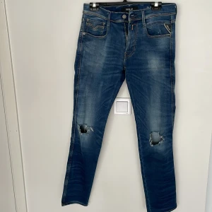 Replay Anbass Hyperflex jeans blå - Blå Replay Anbass Hyperflex jeans med slitningar och hål på båda knäna. Klassisk femficksmodell med snygga detaljer och stretchigt material för extra komfort. Perfekta för dig som gillar en avslappnad och trendig look.
