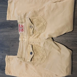 Beiga jeansbyxor Fashion Denim stl 40 - Snygga beiga jeansbyxor från Fashion Denim med coola fickdetaljer och kontrastsömmar. Byxorna har knappar och dragkedja framtill samt fickor med lock och knapp bak. Perfekta för dig som gillar unik stil och detaljer. Det finns lite lösa trådar skriv till mig för fler bilder. Står storlek 40 känns som M men dom är väldigt stretchiga. 