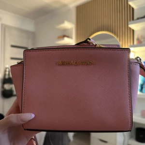 Rosa axelväska från Michael Kors - Snygg rosa axelväska från Michael Kors med guldiga detaljer och stilren design. Väskan är i ett tåligt skinnmaterial och har dragkedja upptill samt ett innerfack. Har några fläckar på sig därför säljs den så billigt 