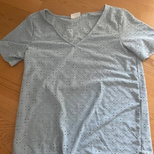 Ljusblå blus med broderi från Vila - Supersöt ljusblå blus från Vila med v-ringning och korta ärmar. Blusen har ett broderat hålmönster över hela tyget som ger en romantisk vibe. Perfekt till jeans eller kjol för en fräsch look.
