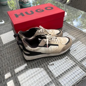Snygga Hugo Boss sneakers, strl 41 - Säljer ett par stilrena sneakers från Hugo Boss (modell HUGO) i storlek 41. Beige och svarta detaljer, klassisk design som funkar både till vardags och lite mer uppklätt. Skorna är använda men i helt okej skick – se bild för detaljer.  Nypris 1599 kr, säljes nu för 200 kr!  Pris: 200 kr Storlek: 41 Märke: Hugo Boss
