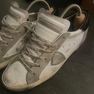 Philippe Model sneakers i vitt och grått - Snygga Philippe Model sneakers i vitt skinn med grå mockadetaljer och svart häl. Skorna har rund tå, snörning med logotyp på skosnörena och en platt sula. Perfekta för dig som gillar en stilren men ändå sportig look.