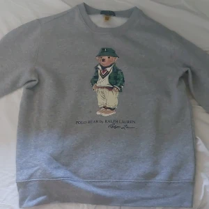 Grå sweatshirt Polo Bear Ralph Lauren - Grå sweatshirt från Polo Ralph Lauren med ikoniska Polo Bear-trycket framtill. Klassisk rund halsringning och långa ärmar. Mjuk insida och relaxed fit, perfekt för chill dagar. Snygg och enkel att matcha med jeans eller joggers.