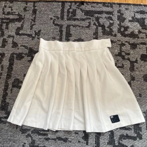 Vit plisserad kjol från Bershka - Vit kort plisserad kjol från Berska med bred linning och dragkedja i sidan. Kjolen har en klassisk tennisstil och är tillverkad i bomullsmix. Perfekt för en sportig och clean look.
