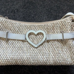 Beige axelväska Suzy Smith hjärtdetalj - Snygg beige axelväska från Suzy Smith i flätad design med vita skinn-detaljer och ett stort hjärta i metall framtill. Väskan har justerbar axelrem och dragkedja upptill. Perfekt för dig som vill ha en söt och trendig accessoar.