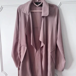 Ljusrosa trenchcoat från Bik Bok - Säljer en ljusrosa trenchcoat från Bik Bok i mjukt material. Jackan har långa ärmar, bred krage och snyggt vattenfallsfall framtill. Den knyts i midjan med bälte och har två stora fickor. Perfekt för vår och höst när du vill ha en chill men stilren look.