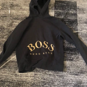 Svart hoodie från Hugo Boss med guldtryck - Svart hoodie från Hugo Boss med stort guldigt BOSS-tryck och HUGO BOSS-text framtill. Klassisk huva med snörning och känguruficka. Långärmad och normal passform, perfekt för en chill och stilren look.