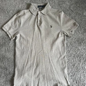 Grå pikétröja Polo Ralph Lauren M - Snygg grå pikétröja från Polo Ralph Lauren i slim fit. Klassisk krage med två knappar och den ikoniska broderade loggan på bröstet. Tillverkad i mjuk bomull, perfekt för en clean och stilren look.