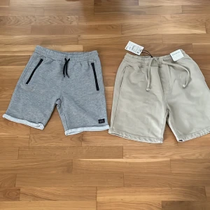 Två par mjuka shorts med snörning - Två par bekväma shorts i mjuk bomull, ett par i ljusgrått med svarta dragkedjor och snörning i midjan, det andra i beige med matchande snörning. Perfekta för chill dagar eller träning. Båda har elastisk midja och praktiska fickor. Den grå är det 158/164 och de beiga är det XS. 60kr för de beiga och 40 kr för de grå.
