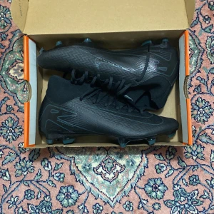 Svarta Nike fotbollsskor med snörning - Snygga helsvarta Nike fotbollsskor med strumpliknande skaft och snörning. Skorna har mönstrad ovandel i syntetmaterial och dobbar för gräsplan. Perfekta för dig som vill ha en clean look på planen och maximal kontroll över bollen.