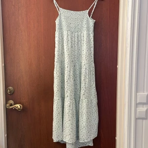 Ljusblommig långklänning Basic Needs - Barnklänning strl 152 . Superfin långklänning med smala axelband från Basic Needs. Ljus botten med små blommor i lila och grönt, smock upptill och luftig kjol. Perfekt för varma dagar och somrig stil.
