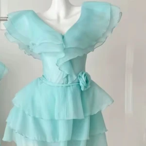 Ljusblå volangklänning med rosdetalj - Superfin ljusblå kortklänning med flera lager volanger och v-ringad omlottfront. Klänningen har en markerad midja med ett bälte och en söt tygros på sidan. Tillverkad i ett lätt, flowy material som ger en drömmig känsla.