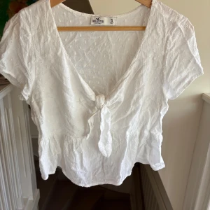 Vit blus med knyt från Hollister - Supersöt vit blus från Hollister med korta ärmar, v-ringning och knyt framtill. Blusen har ett diskret prickigt strukturmönster och volangkant nedtill. Perfekt för en fräsch och somrig look!