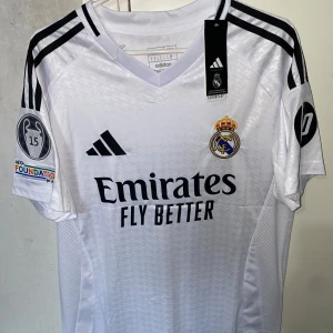 Real Madrid Mbappé 9 matchtröja Adidas - HELT NY Real Madrid fotbollströja med Mbappé och nummer 9 på ryggen. Vit med svarta detaljer, klubbmärke, Adidas-logga och sponsortryck. Korta ärmar med svarta ränder och patchar på båda ärmarna. Tillverkad i lätt, ventilerande material. STORLEK: S.      OM PRISFÖRSLAG MEDDELA MIG