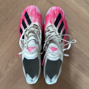 Adidas fotbollsskor i vitt och rosa - Snygga Adidas fotbollsskor med vit ovandel, rosa detaljer och svarta ränder på sidorna. Skorna har snörning och är tillverkade i syntetmaterial för en lätt känsla på planen. Perfekta för dig som vill sticka ut med färg och stil under matchen. Använda fåtal gånger