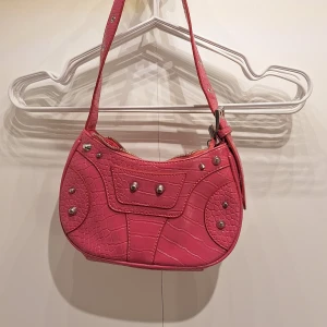 Rosa axelväska - Cool rosa axelväska med croco-mönster och silvriga nitar. Väskan har dragkedja upptill, en ficka med dragkedja på baksidan och snygga detaljer framtill. Perfekt storlek för mobilen och det viktigaste. Fodrad insida i matchande rosa. Köpt på Ullared