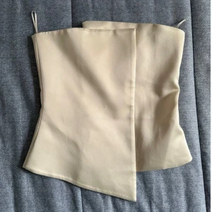 Beige asymmetrisk tubtopp i satin - Säljer en beige tubtopp med asymmetrisk skärning och stilren design. Toppen är axelbandslös och har en lätt glansig satinfinish som ger en lyxig känsla. Perfekt för dig som gillar minimalistiska och trendiga plagg. 