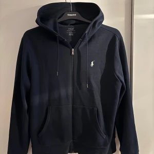 Mörkblå hoodie från Polo Ralph Lauren - Svart hoodie från Polo Ralph Lauren med dragkedja, huva och snörning. Klassisk broderad logga på bröstet och två stora fickor framtill. Skön passform och tillverkad i mjuk bomull, perfekt för chill dagar.