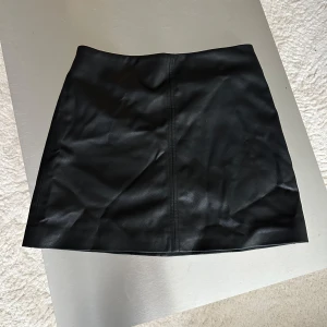 Svart skinnkjol från H&M, storlek 38 - Säljer en svart kort skinnkjol från H&M i storlek 38. Kjolen har en clean look med söm framtill och dragkedja bak 💕
