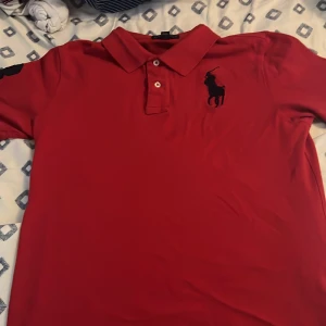Röd pikétröja från Ralph Lauren - Säljer en klassisk röd pikétröja från Ralph Lauren som är köpt från NK för 1200 med svart broderad logga på bröstet och en stor svart 3:a på ärmen. Tröjan passar dig som bär storleke xs skulle jag säga, Tröjan har krage, två knappar och är kortärmad. Perfekt för en clean och sportig look. 
