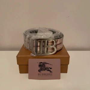 Rutig Burberry bälte med B-spänne - Snyggt bälte från Burberry med klassiskt rutigt mönster i beige, grått och rött. Stort silvrigt B-spänne framtill som ger en lyxig känsla. Perfekt accessoar för att lyfta din outfit med en ikonisk touch.