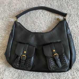 INTRESSEANMÄLAN - Svart axelväska i skinnimitation med två framfickor med spännen och flätad detalj. Väskan har guldfärgade metalldetaljer och dragkedja både upptill och på baksidan. Perfekt storlek för vardagens essentials och snygg att bära över axeln. OBS: INTE RIKTIGA PRISET