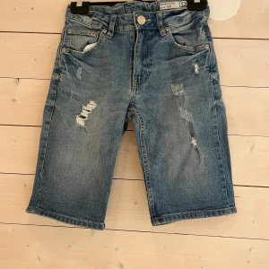 Blå slitna jeansshorts Shorty Star - Snygga blå jeansshorts från Shorty Star i storlek 150. Modellen är knälång med klassisk femficksdesign, slitna detaljer framtill och normal passform. Perfekta för en chill sommarlook med t-shirt eller hoodie.