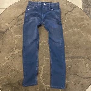 Blå skinny jeans från New Denim - Snygga blå skinny jeans från New Denim med slitna detaljer på låren och klassiska fem fickor. Jeansen har vita kontrastsömmar och en smal passform som sitter tajt längs benen. Perfekta för dig som gillar en modern och enkel look.