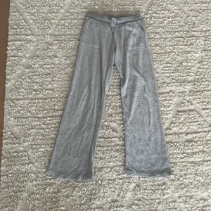 Grå mjukisbyxor Juicy Couture - Grå mjukisbyxor från Juicy Couture med broderad JC-logga på ena benet. Byxorna har dragsko i midjan, fickor fram och bak samt en loose passform med raka ben. Perfekta för chill dagar eller när du vill vara extra bekväm.