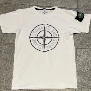 Stone Island tröja - Vit t-shirt från Stone Island med stor broderad logga på bröstet och klassisk patch på ärmen. T-shirten har rund hals och korta ärmar, tillverkad i mjuk bomull. Perfekt för dig som gillar streetwear och vill sticka ut med en ikonisk look.