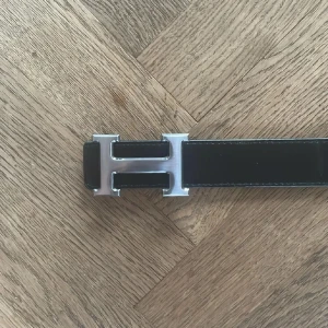 H bälte vändbart - Säljer ett H bälte med ikoniskt H-spänne i silverfärgad metall. Ena sidan är svart och den andra är blå, båda i slätt skinn med kontrastsömmar. Perfekt accessoar för att lyfta din outfit med en lyxig touch och klassisk design.