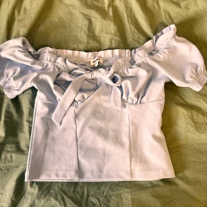 Ljusblå offshoulder topp med knyt - Supersöt ljusblå offshoulder topp från bubbleroom med puffiga korta ärmar och knyt framtill. Toppen har en feminin och romantisk vibe, perfekt till jeans eller kjol. Mjuk och skön i materialet, med fina rynkade detaljer vid axlarna.