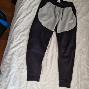 Nike mjukisbyxor svart/grå S - Nike mjukisbyxor i svart och grått med snörning i midjan och logga på vänster sida. Byxorna har ribbade muddar vid benslut och är tillverkade i mjukt material för maximal komfort. Perfekta för chill eller träning. De har ett litet märke på vänsterbenet (se bild 4) men det syns knappt på avstånd