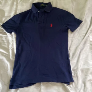 Marinblå pikétröja Polo Ralph Lauren - Snygg marinblå pikétröja från Polo Ralph Lauren i slim fit-modell. Klassisk krage, korta ärmar och röd broderad logga på bröstet. Tillverkad i bomull, perfekt för en clean och stilren look.