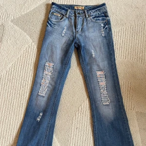 Blå slitna lågmidjade bootcut jeans med detaljer - Säljer ett par blå jeans med bootcut passform och slitna detaljer framtill och baktill. Jeansen har klassisk femficksdesign, låg midja och coola slitningar på både ben och bakfickor. Perfekta för dig som gillar en avslappnad och trendig stil.