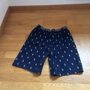 Mörkblå shorts Polo Ralph Lauren - Snygga mörkblå shorts från Polo Ralph Lauren med klassiskt vitt logomönster över hela tyget. Resår i midjan med tydlig logga. Perfekta för chill dagar eller när du vill ha en clean och stilren look.