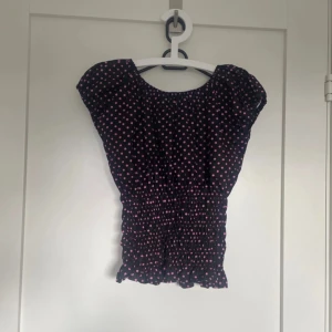 Svart prickig blus med resår - Supersöt svart blus med rosa prickar och smockad midja. Toppen har korta puffärmar och en rundad halsringning. Den är lätt och luftig, perfekt för varma dagar. Den smockade delen ger en snygg passform och volangkant nertill gör den extra gullig.