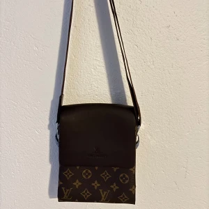 brun axelväska  - Snygg brun axelväska med klassiskt monogrammönster och justerbar rem. Väskan har lock i mörkbrunt skinn och flera fack med dragkedja inuti. Perfekt för dig som vill ha en stilren och ikonisk accessoar.