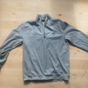 Grå half zip tröja från Polo Ralph Lauren - Snygg grå långärmad tröja från Polo Ralph Lauren med half zip och broderad logga på bröstet. Tröjan har en hög krage och är tillverkad i mjuk bomull, perfekt för lager-på-lager-stil. Enkel och stilren design som funkar året runt.