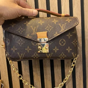  monogram handväska - Snygg handväska i klassiskt brunt monogrammönster. Väskan har guldfärgade metalldetaljer, kedjerem och justerbar axelrem i skinn. Invändigt finns dragkedjefack och flera fack för småsaker. Perfekt för dig som vill ha en ikonisk accessoar.